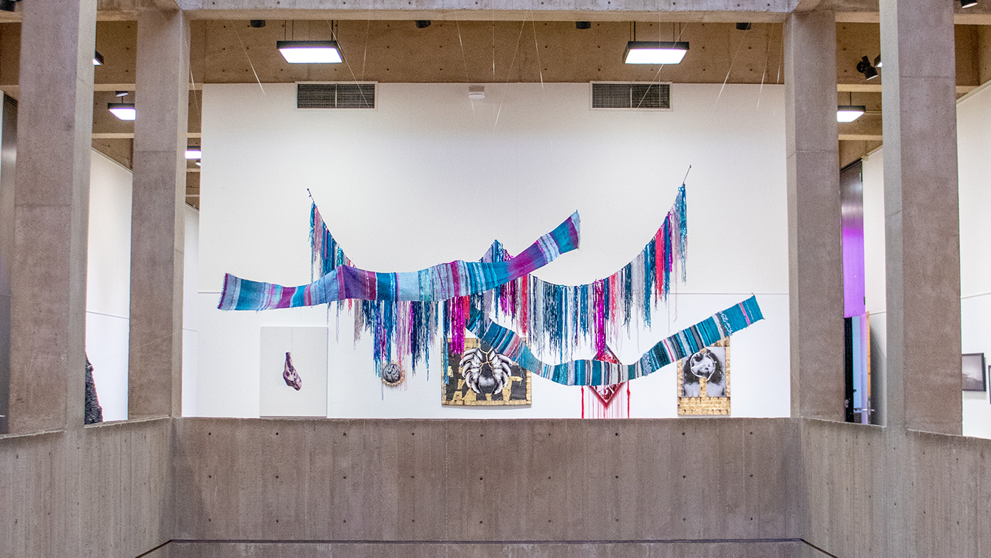 A large weaving by Amy Huser hangs in the McKnight Art Center Atrium in front of three two-dimensional works of art by Sean Heaton and Lily Guillen.