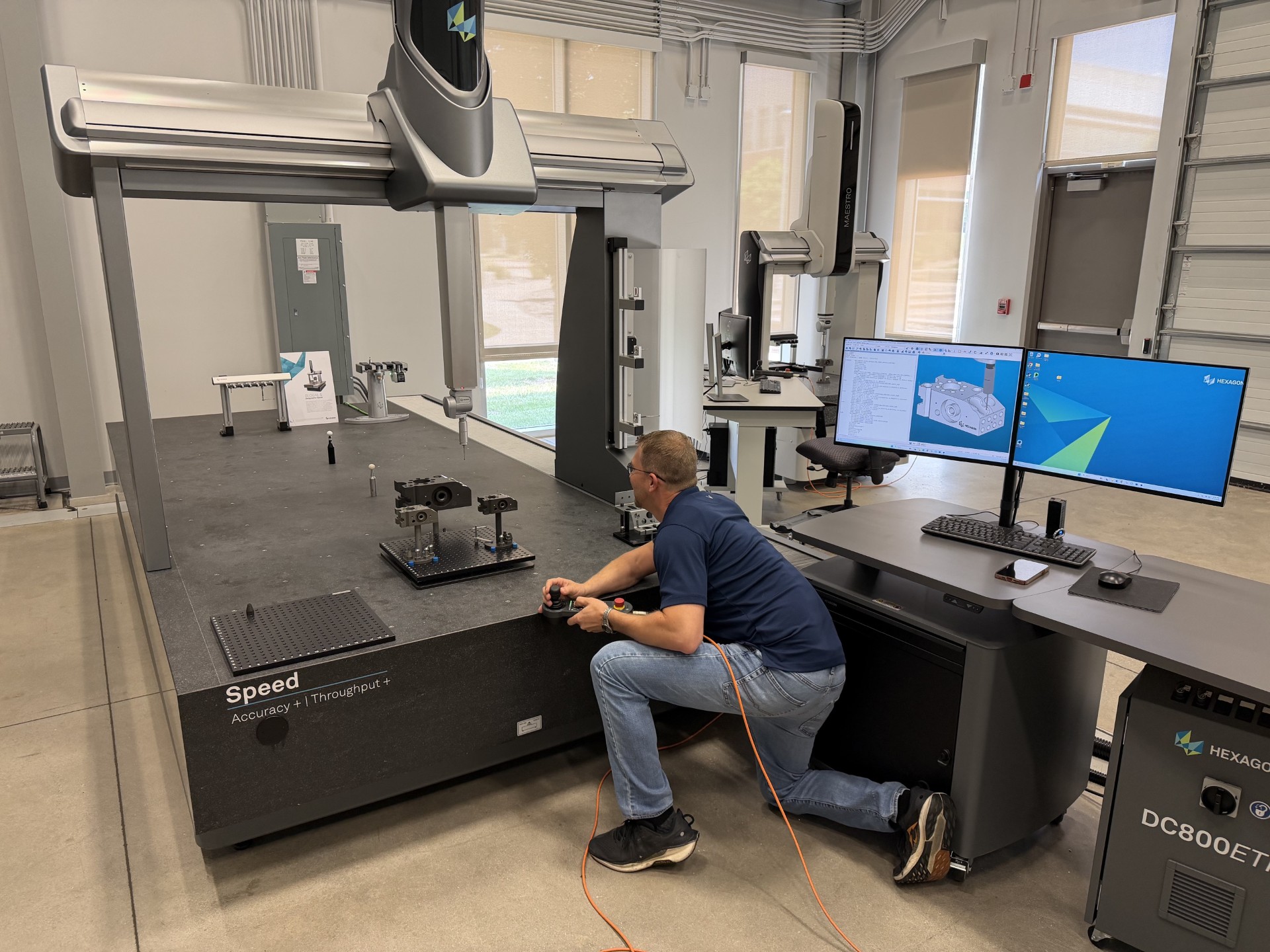The image shows a person working in a modern industrial metrology lab. The individual is kneeling beside a large coordinate measuring machine (CMM) with the brand “Hexagon” visible on the equipment. The machine has a wide granite base labeled “Speed – Accuracy + Throughput,” and several metal components and fixtures are arranged on its surface for measurement. The person is holding a controller connected by an orange cable, appearing to adjust or operate the CMM. Nearby is a workstation with dual monitors displaying CAD models and measurement software. The workspace includes a keyboard, mouse, and additional computer equipment on a sleek, dark workstation cabinet labeled “DC800ET.” The room itself has high ceilings, exposed conduit, large windows, and polished concrete floors, giving it a clean, industrial, and high-tech environment. Multiple pieces of precision equipment and workstations are positioned around the room, indicating a professional engineering or manufacturing measurement facility.